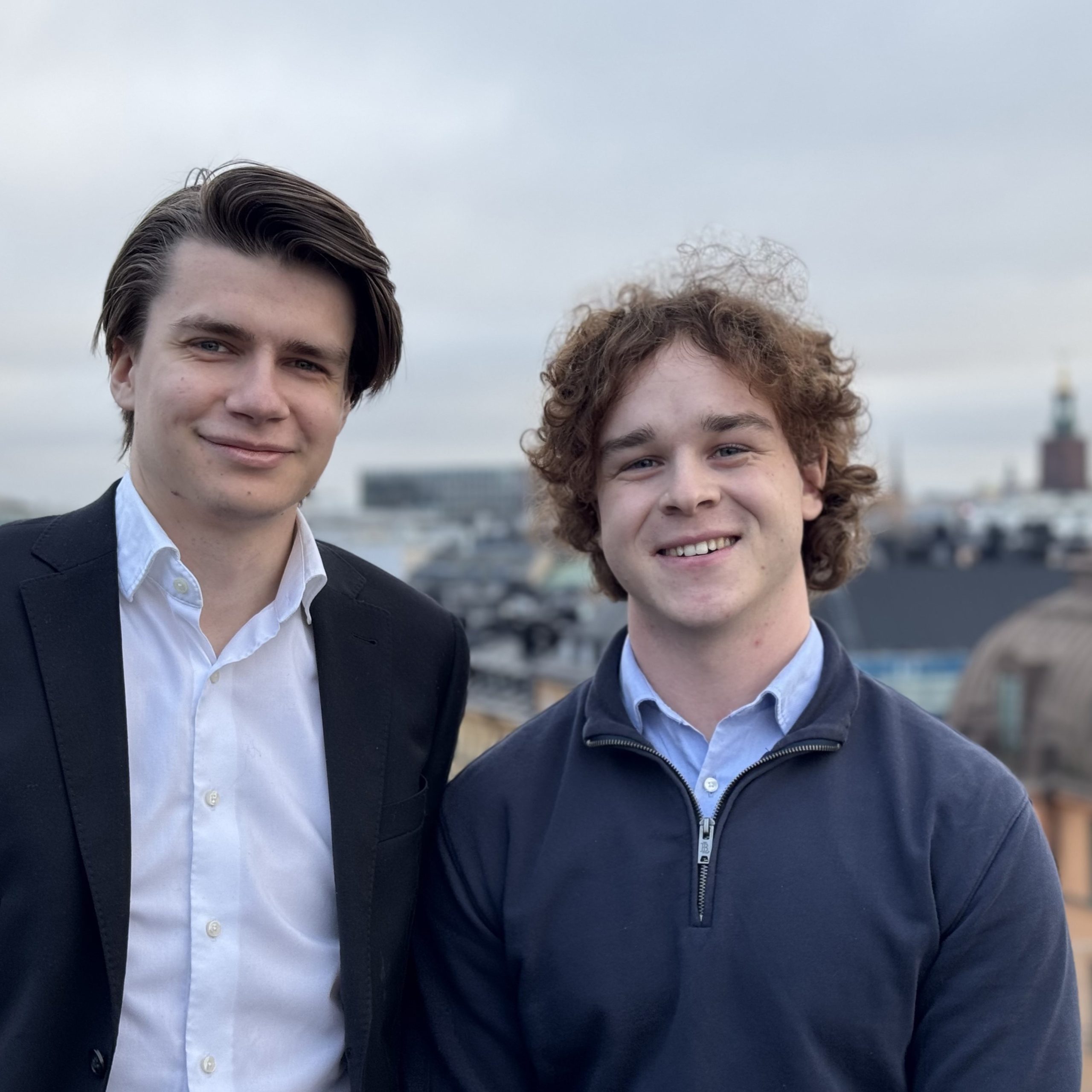 Två personer står utomhus med en stadsmiljö i bakgrunden.