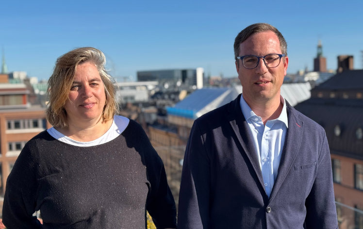 2025-04-09 Ruth o Agustin Två personer står på ett tak med stadslandskap och blå himmel i bakgrunden.