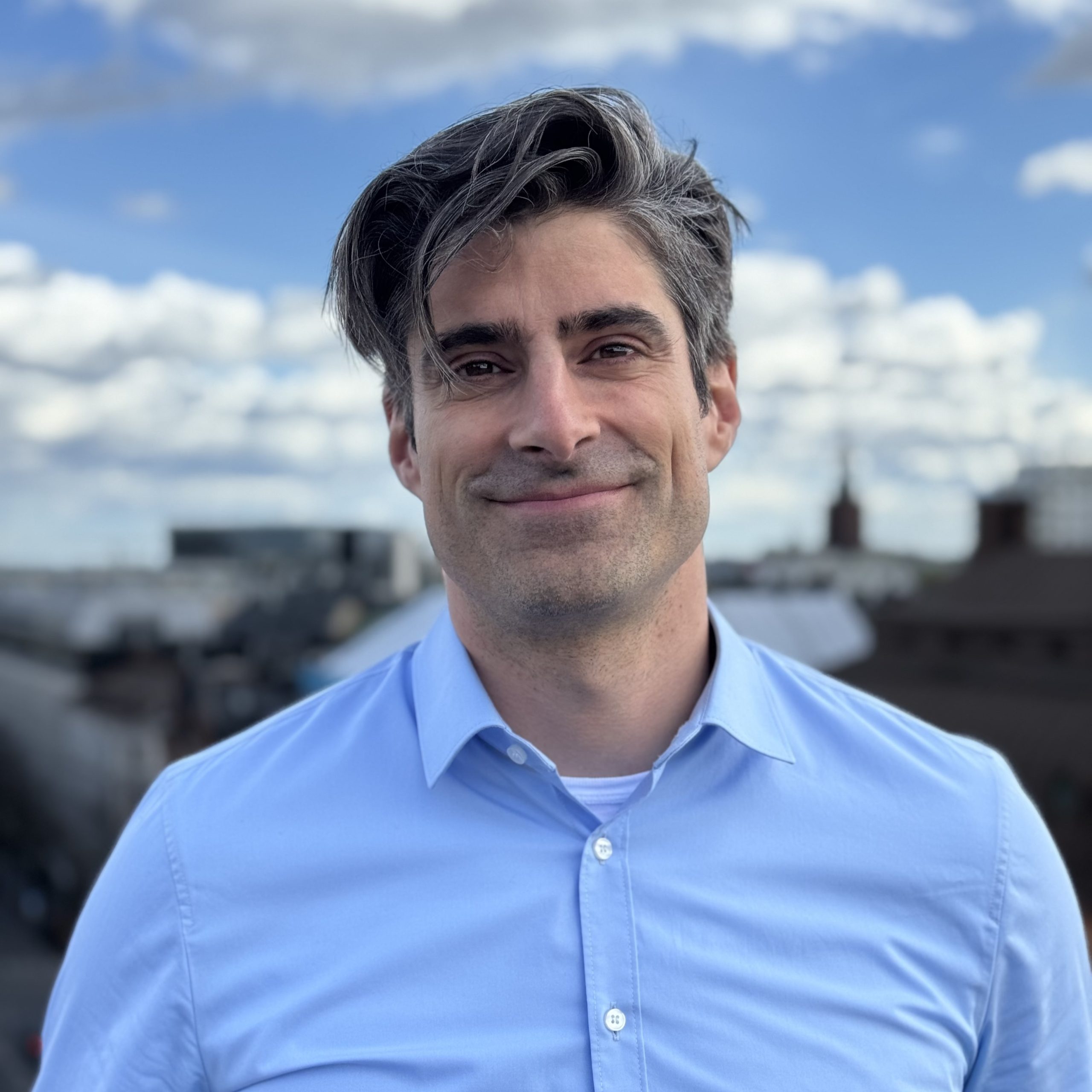 Person står utomhus med byggnader och himmel i bakgrunden.