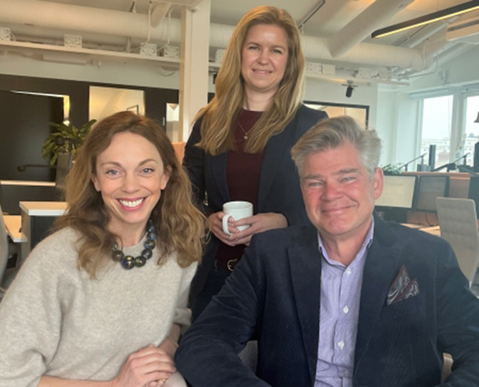 En bild av två sittandes personer med en person som står bakom dem, samtliga personer ler mot kameran.