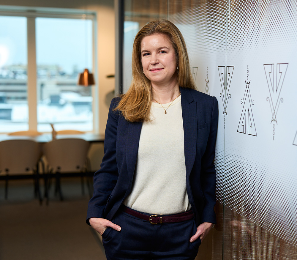 Porträtt av Carolin Sundkvist som står i kontorsmiljö, lutad mot en vägg med Groth & Co:s logotyp