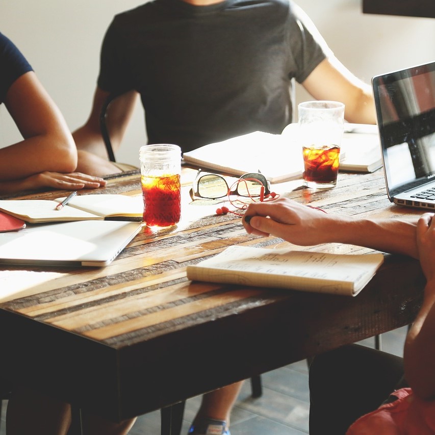 Personer som sitter runt ett bord med anteckningsblock, laptop och dryck under ett informellt möte.