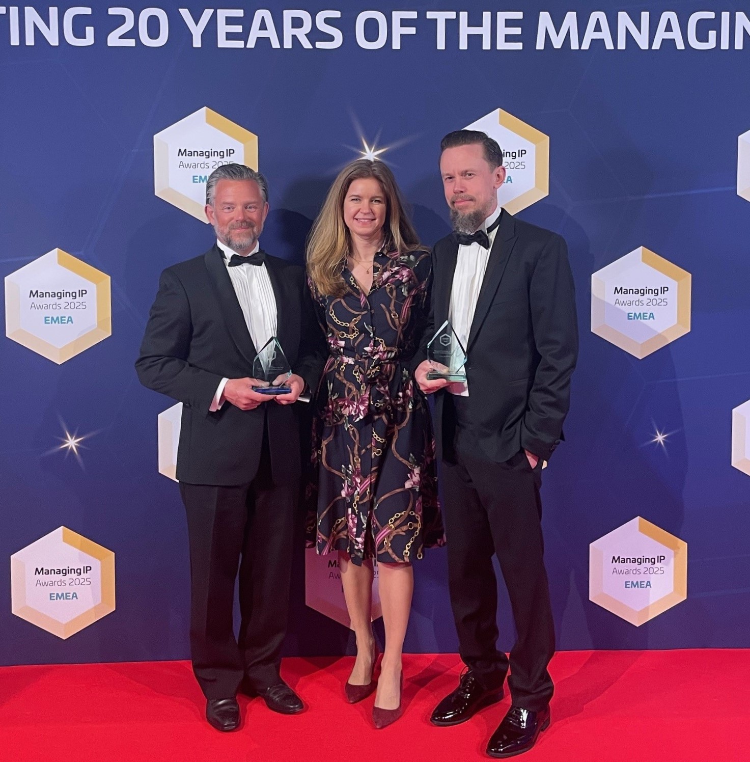 Tre personer på galamiddag poserar på röd matta med prisstatyetter.